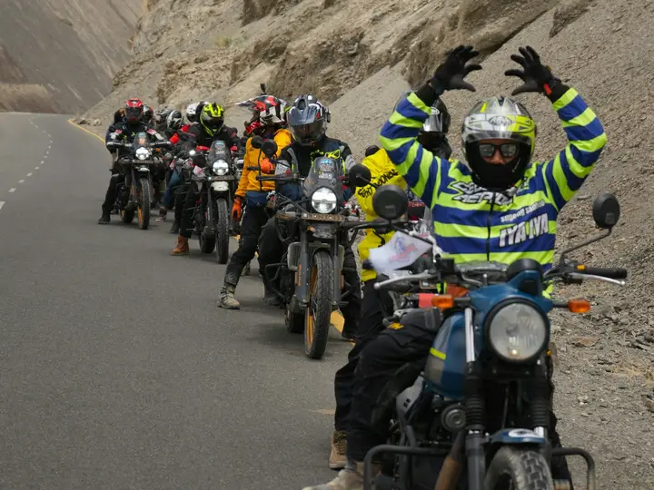 A long line of riders on a mountain road, one waving with both arms raised